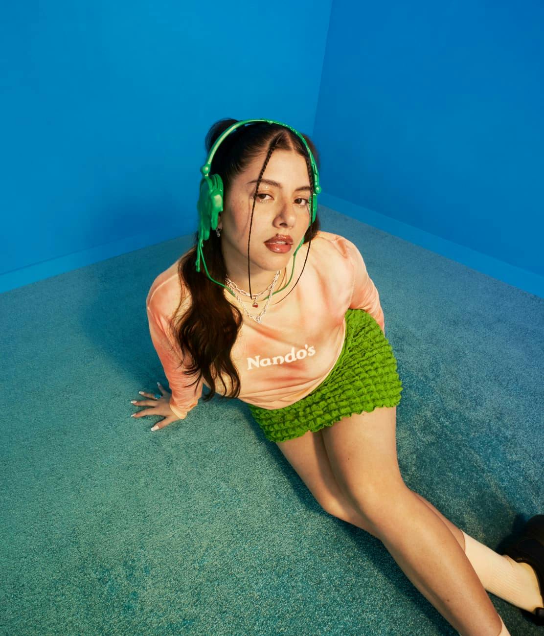 High-angle shot of a young woman with braided hair, wearing a peach Nando's tie-dye long-sleeve top, a bright green textured mini skirt, and large green headphones. She is seated on a blue carpeted floor against a blue wall, looking directly up at the camera with a neutral expression.