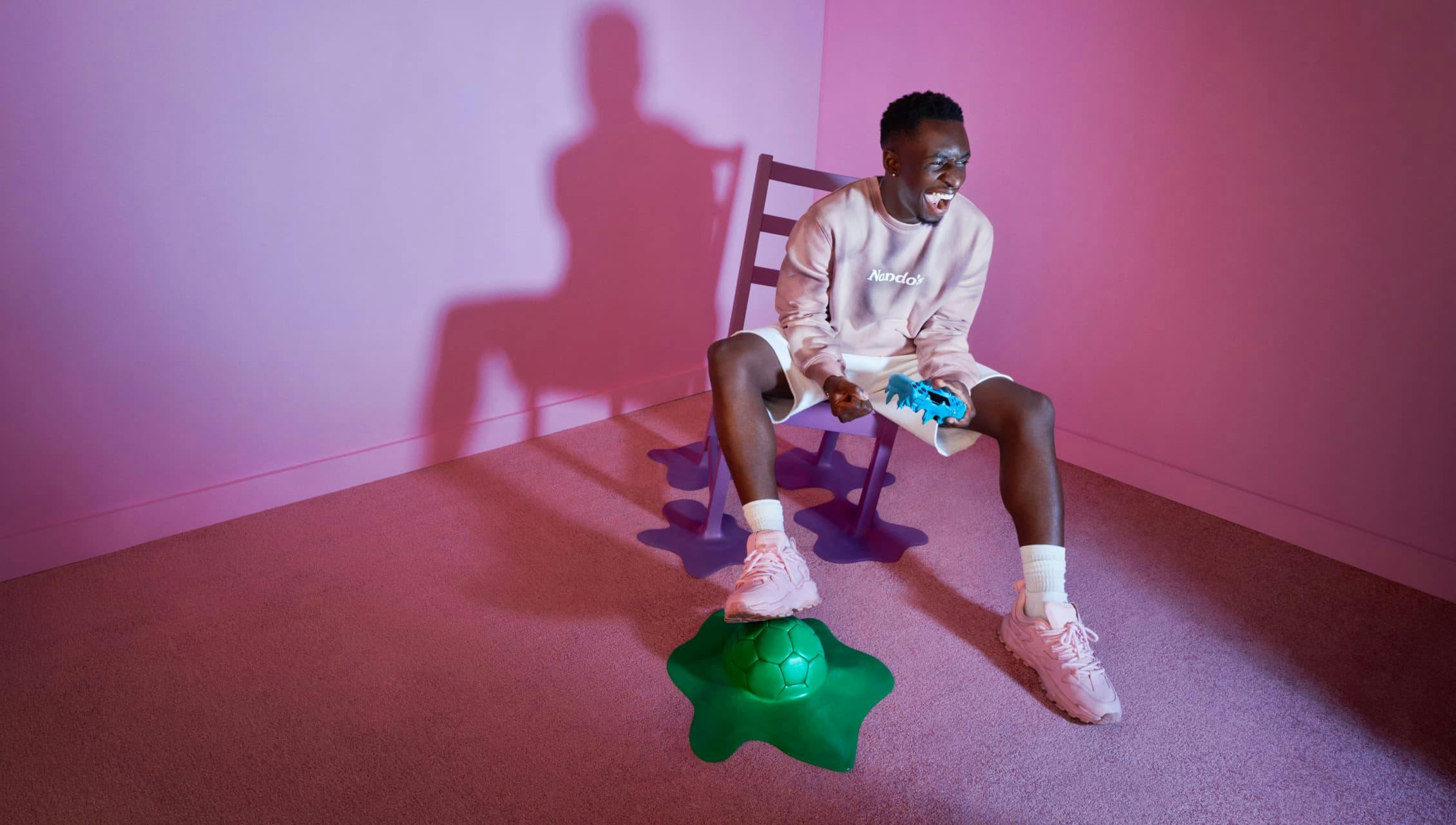 A young man wearing a light pink Nando's sweatshirt and white shorts, enthusiastically playing a video game with a blue controller. He's seated on a purple chair in a room with pink walls and carpet, his right foot resting on a green, splat-shaped soccer ball.