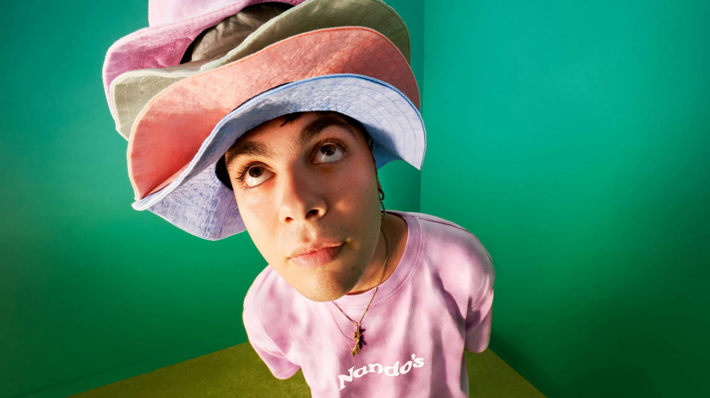 A high-angle, slightly distorted shot of a young person wearing a pink Nando's t-shirt and a precarious stack of five different colored bucket hats (pink, grey, coral, light blue). They are looking up with a surprised or curious expression against a vibrant green backdrop.