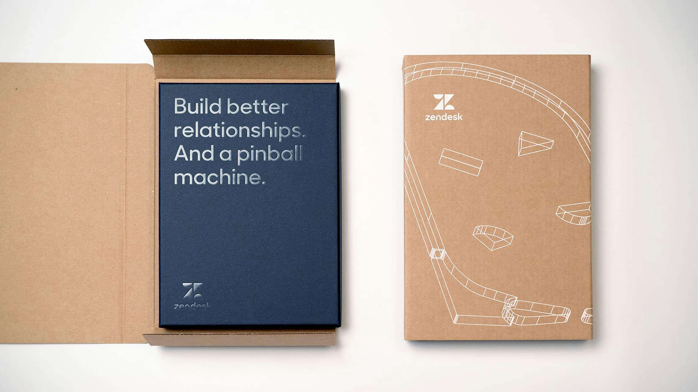 A flat lay image of Zendesk-branded packaging. On the left is an open cardboard mailer box revealing a premium dark blue box inside. The blue box has silver foil text reading 'Build better relationships. And a pinball machine.' On the right is a matching cardboard sleeve with the Zendesk logo and a white line art illustration of a pinball machine.