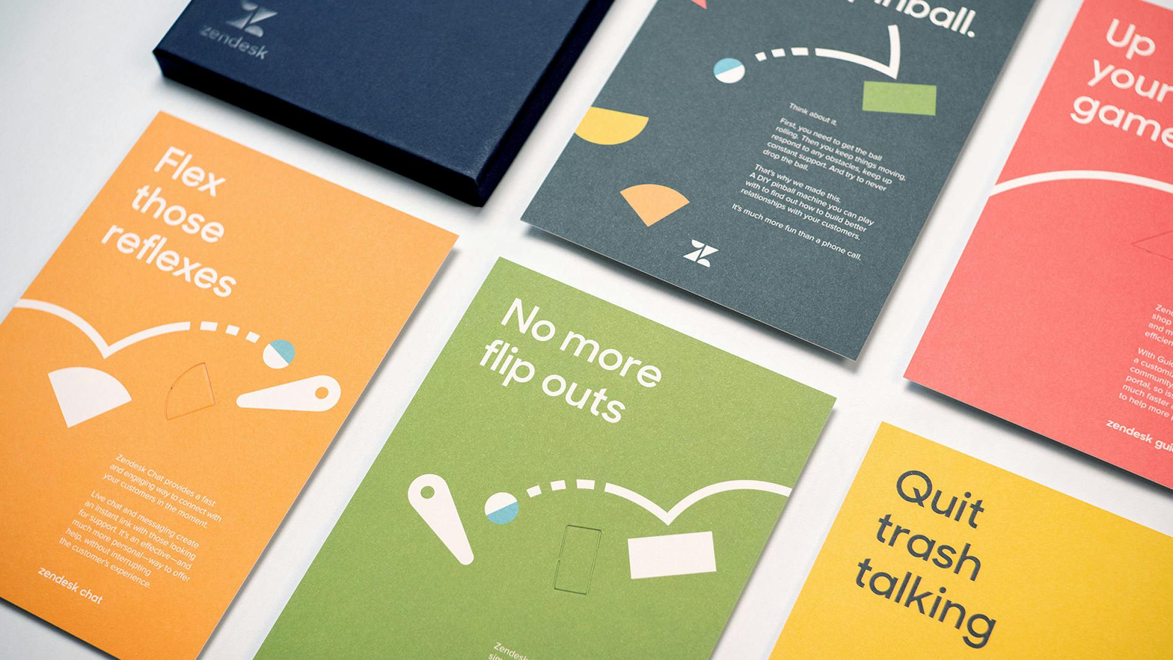 A flat lay of various colorful printed cards from a Zendesk marketing kit. Each card has a different color (orange, green, yellow, red, dark grey) and features a pinball-themed headline and graphics, connecting pinball concepts to Zendesk product benefits. A corner of a dark blue Zendesk box is also visible.