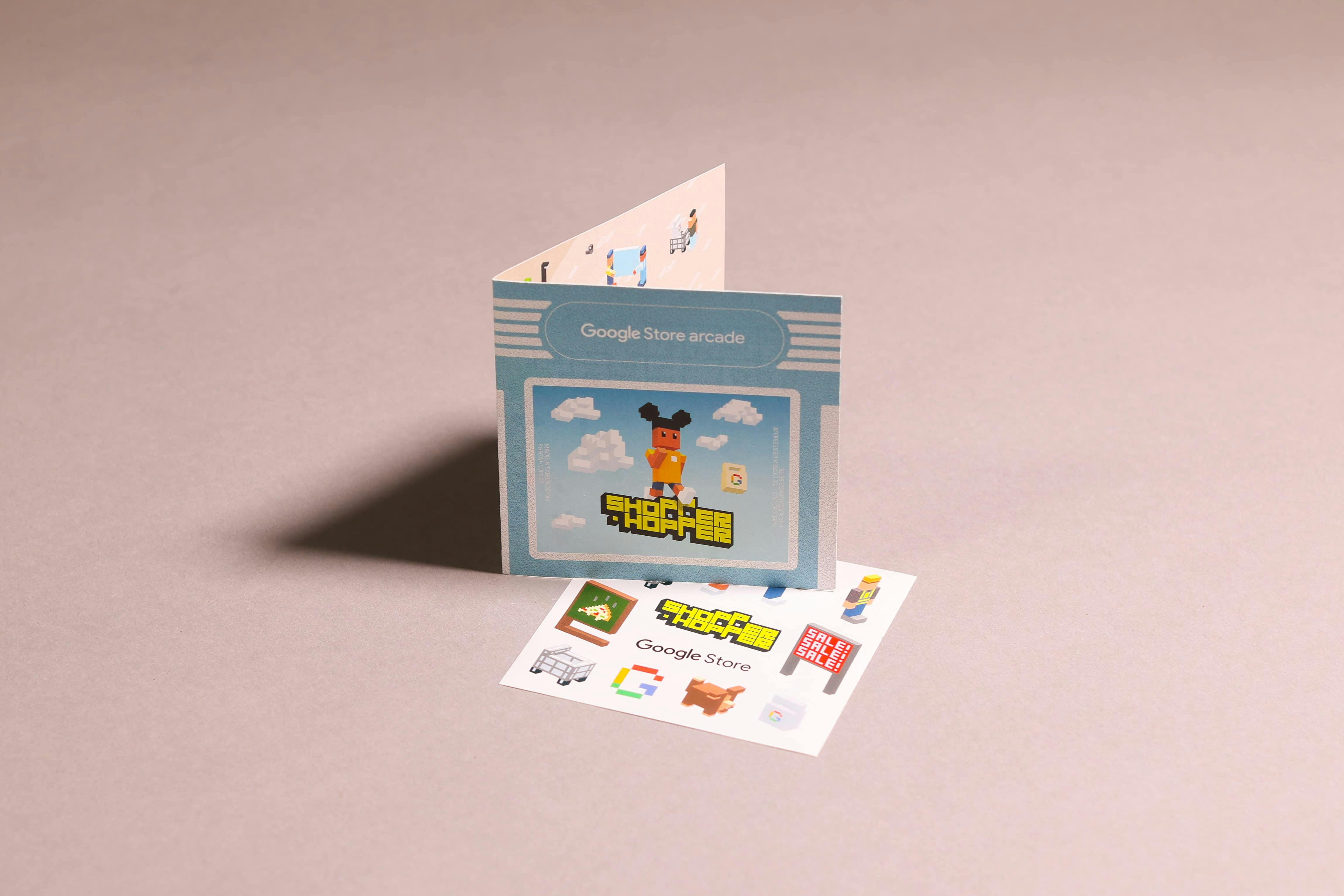 A still life of 'Google Store arcade' promotional materials. A folded card stands upright, showing the 'Shopper Hopper' logo and main character. In front of it lies a sticker sheet with various voxel art images from the game, including a shopping cart, sale sign, and the Google 'G' logo.