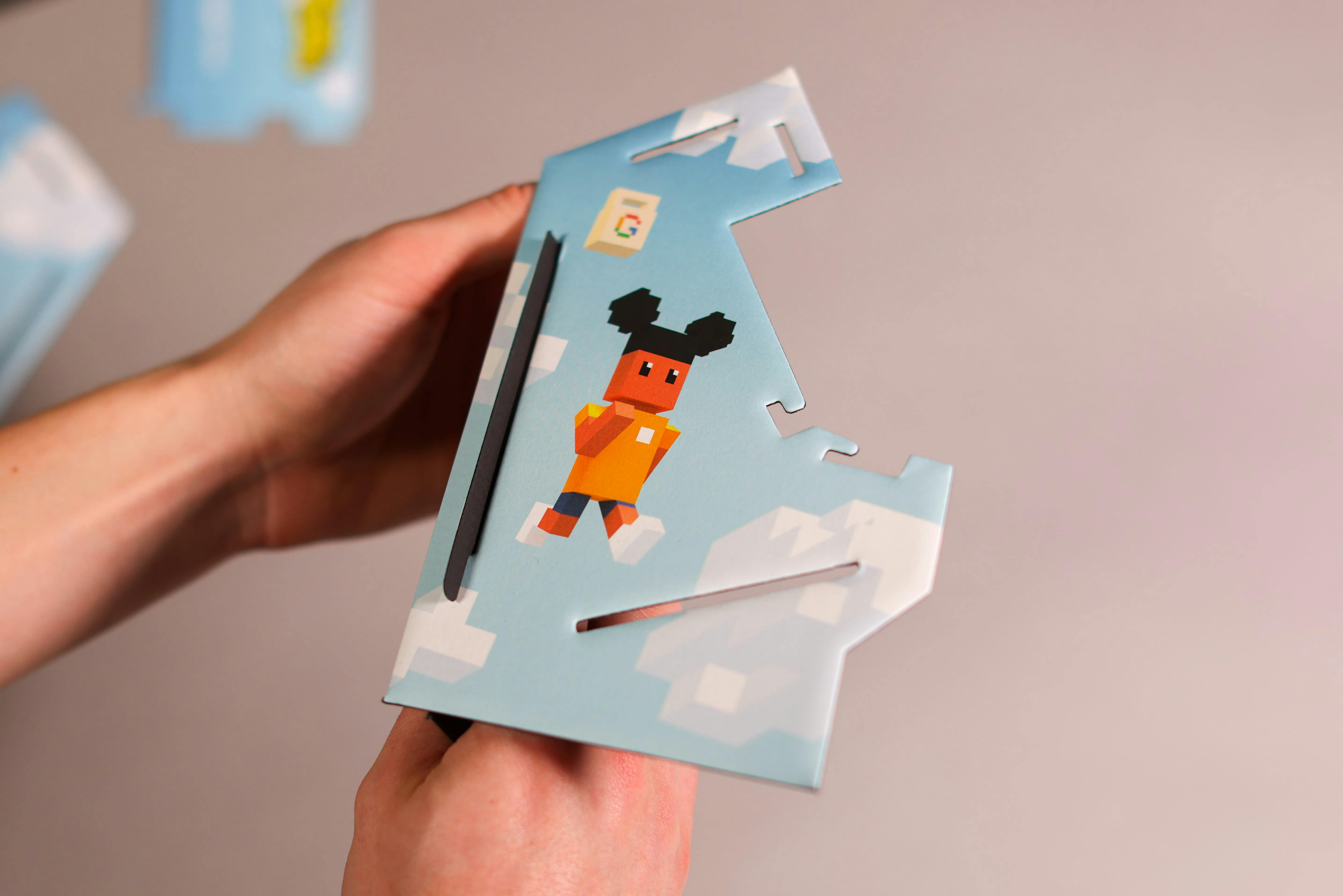 Close-up of a person's hands holding a flat, die-cut piece of cardboard. The piece is printed with artwork from the 'Shopper Hopper' game, featuring the main character in voxel style, a Google shopping bag, and a background of a blue sky with pixelated clouds. The piece has notches and slots, indicating it's part of a buildable model.