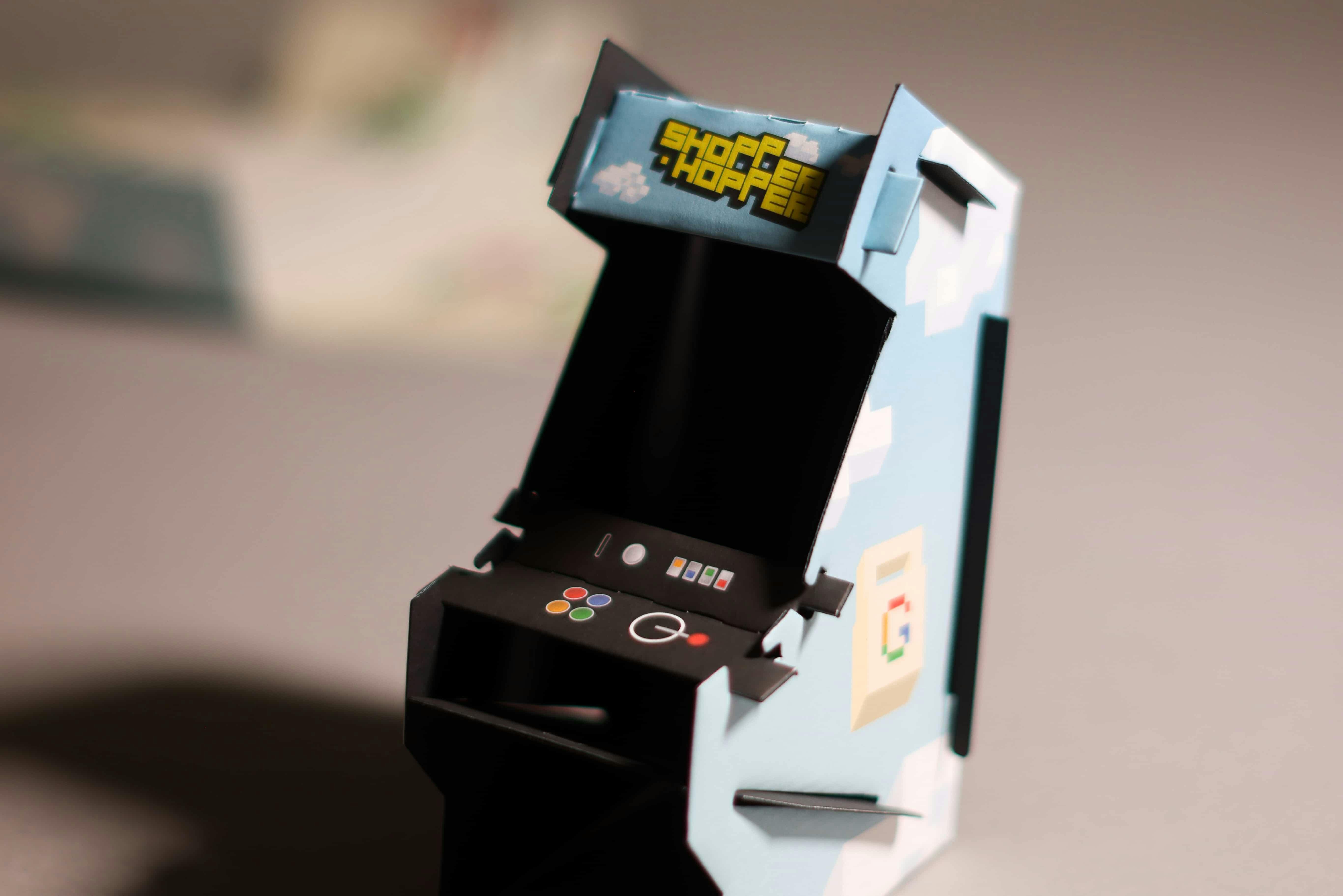 A small, self-assembled cardboard model of a classic arcade game cabinet. The sides are light blue with voxel-art clouds, while the marquee at the top features the 'Shopper Hopper' logo. The black control panel has printed-on buttons in Google colors (red, yellow, green, blue).