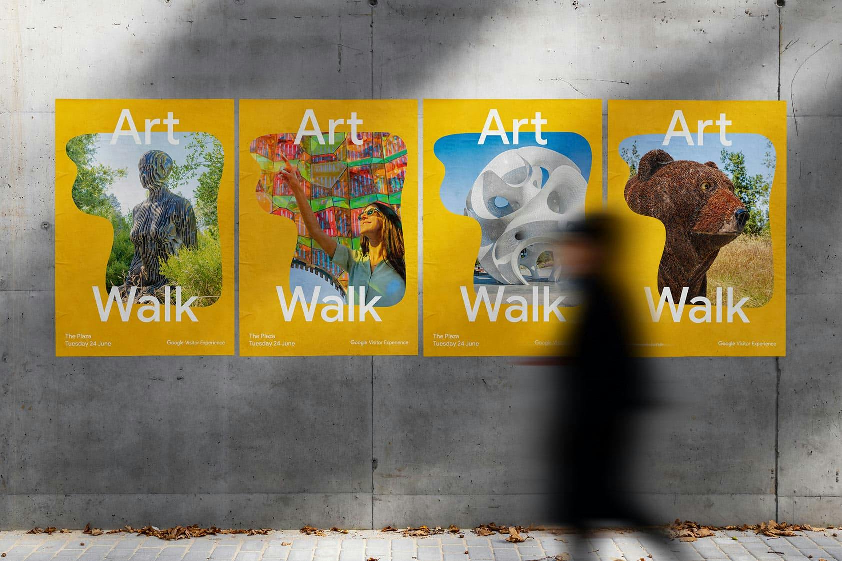 Four bright yellow posters on a concrete wall promoting the Google Visitor Experience Art Walk, each featuring a different sculpture or artwork including a humanoid figure, colorful canopy, abstract white structure, and a bear sculpture, with a blurred figure walking by.