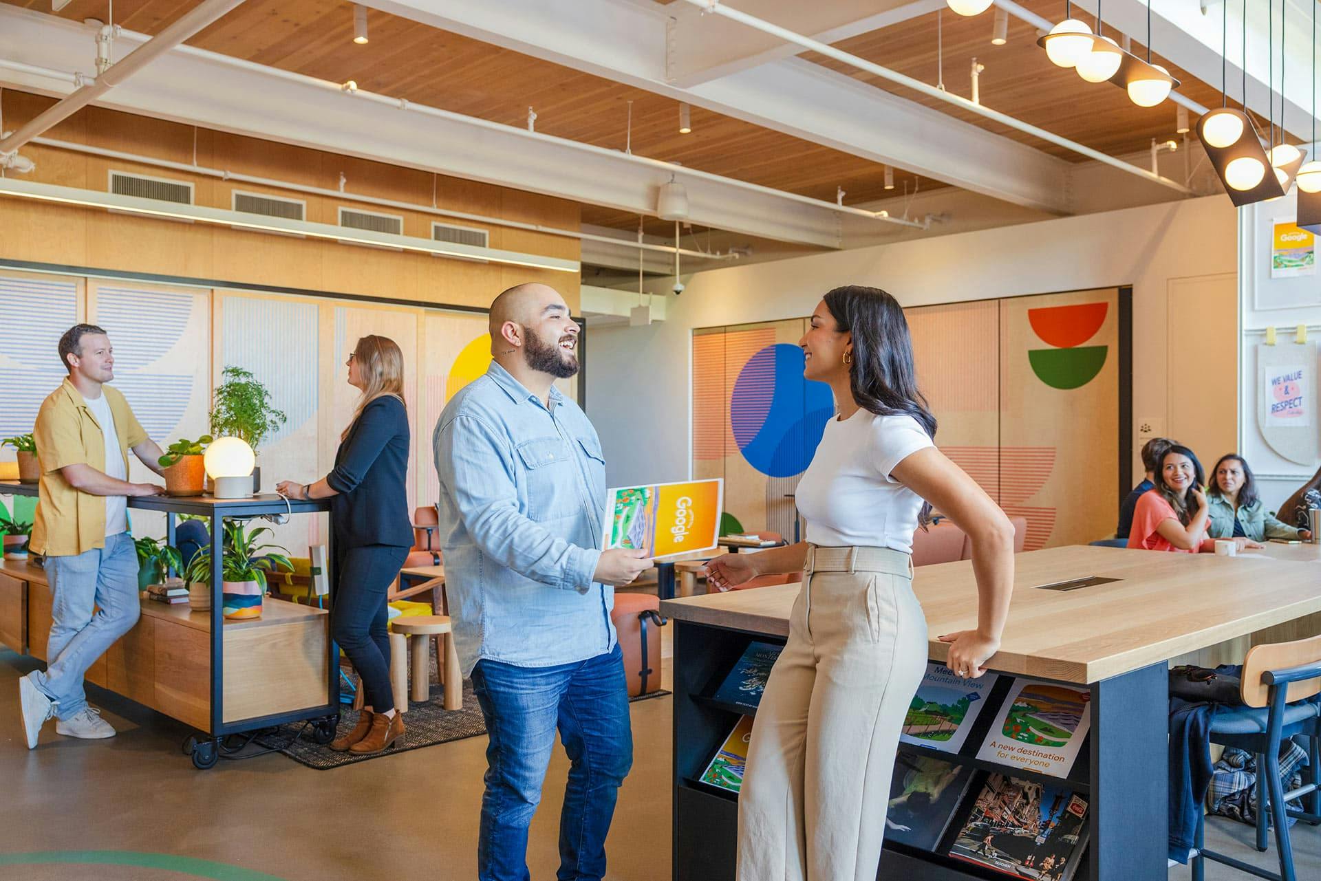 A vibrant scene inside a Google facility, showing a man cheerfully handing a colorful Google brochure to a woman who is smiling and leaning on a high counter. Other people are conversing in the background in a well-lit, modern space with wooden accents and colorful geometric wall art.