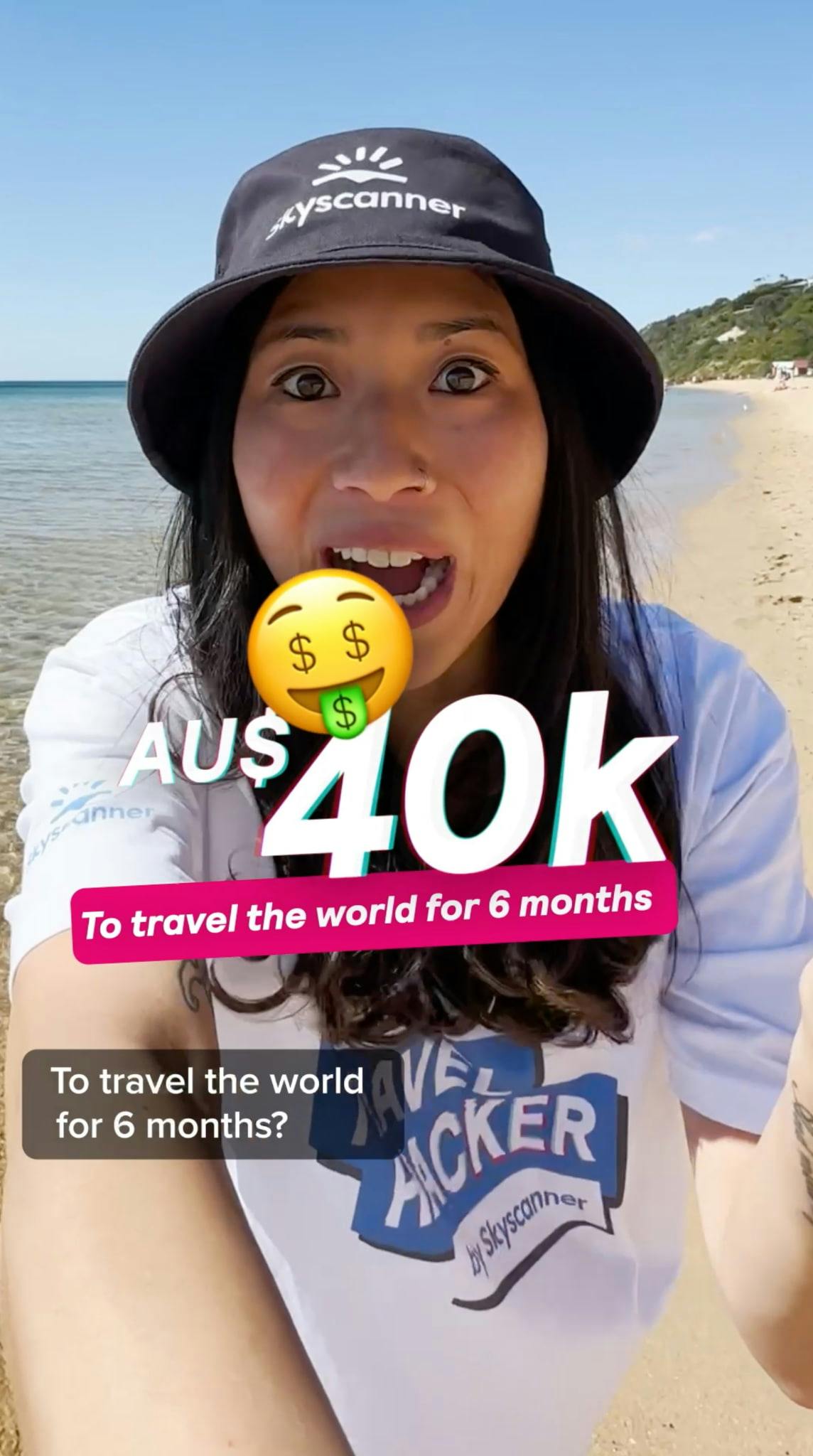 Close-up selfie-style shot of a woman on a sunny beach, wearing a black Skyscanner bucket hat and a white t-shirt with the 'ULTIMATE TRAVEL HACKER by Skyscanner' logo. She has an excited expression with her mouth open. Large text overlays read 'AU$ 40K' with a money-mouth emoji, and below it, 'To travel the world for 6 months' on a pink banner. Subtitles at the bottom reiterate: 'To travel the world for 6 months?