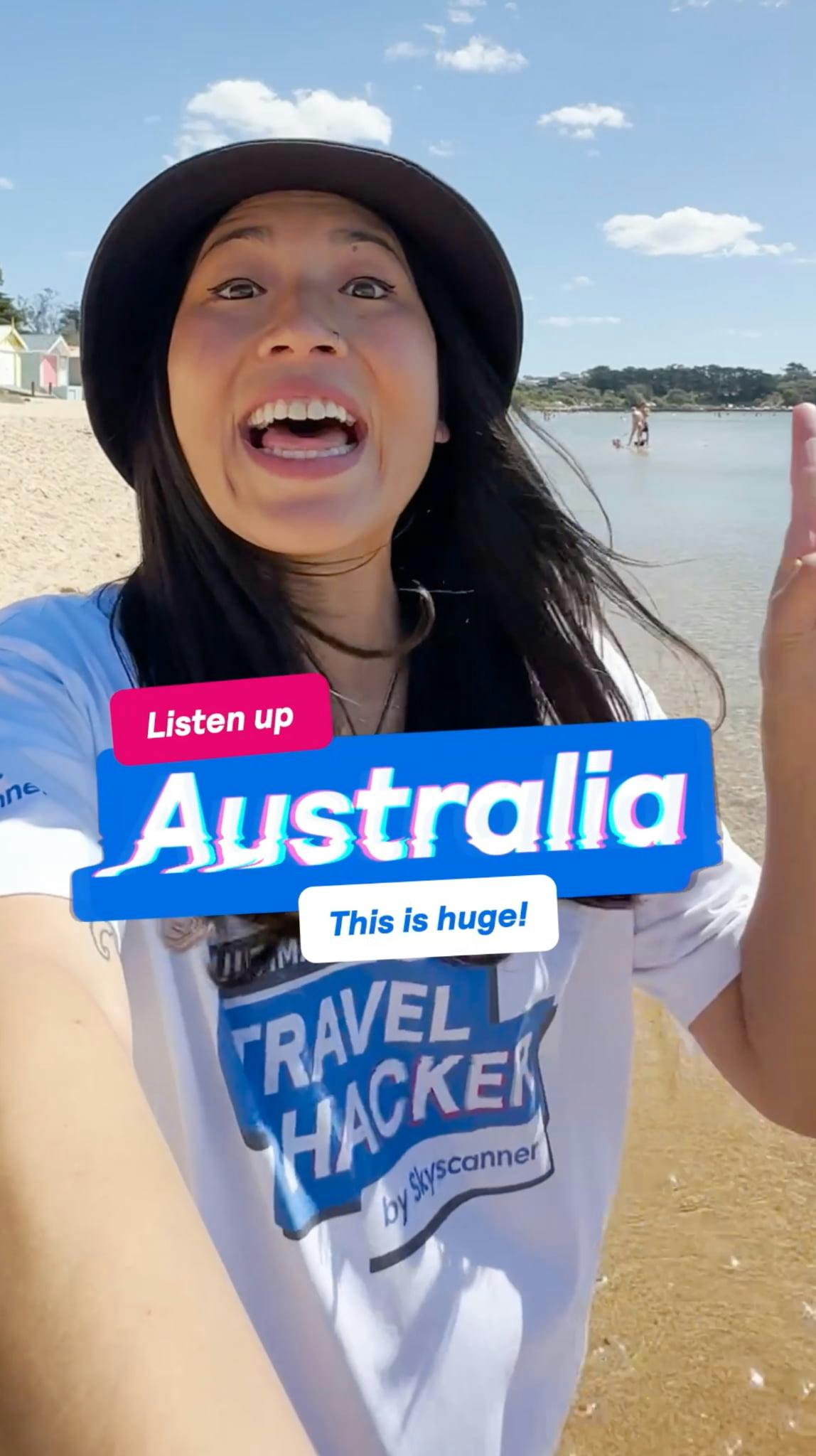 Selfie-style shot of an excited woman on a sunny beach, wearing a black bucket hat and a white t-shirt with the 'ULTIMATE TRAVEL HACKER by Skyscanner' logo. She is pointing off-camera with an open mouth. Text overlays read 'Listen up' on a pink banner, 'Australia' in large, bold white text with a blue glitch effect background, and 'This is huge!' on a white banner.