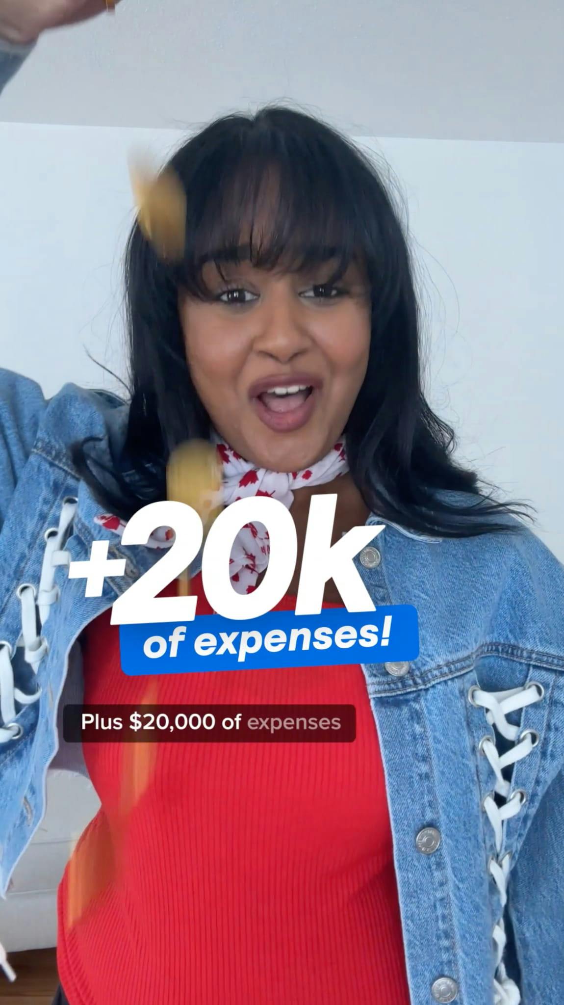 An excited woman with dark hair and bangs, wearing a red top, a denim jacket with white lace-up details, and a white and red patterned neckerchief, looks at the camera with her mouth open. She is tossing what appears to be gold confetti or glitter. Large text overlays read '+20K' in white, with 'of expenses!' on a blue banner below it. A smaller black subtitle banner reads 'Plus $20,000 of expenses'.