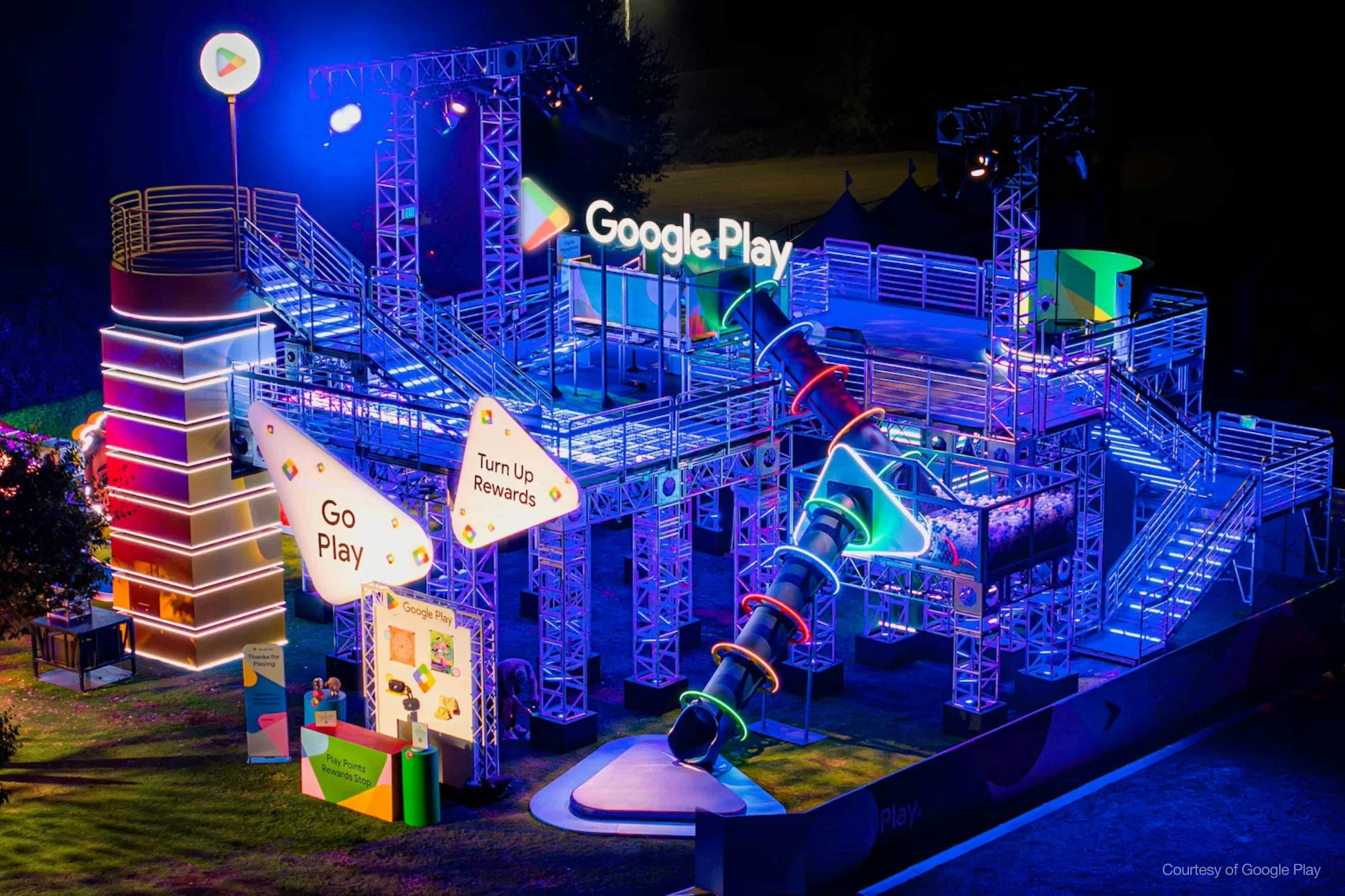 Nighttime shot of a large, brightly lit Google Play interactive event structure. The installation includes a multi-story metal truss framework with stairs, platforms, a large enclosed tube slide adorned with colorful neon rings, and various illuminated Google Play logos and signage like 'Go Play' and 'Turn Up Rewards.' The scene is bathed in vibrant blue, pink, and green lights. 'Courtesy of Google Play' is watermarked in the bottom right.