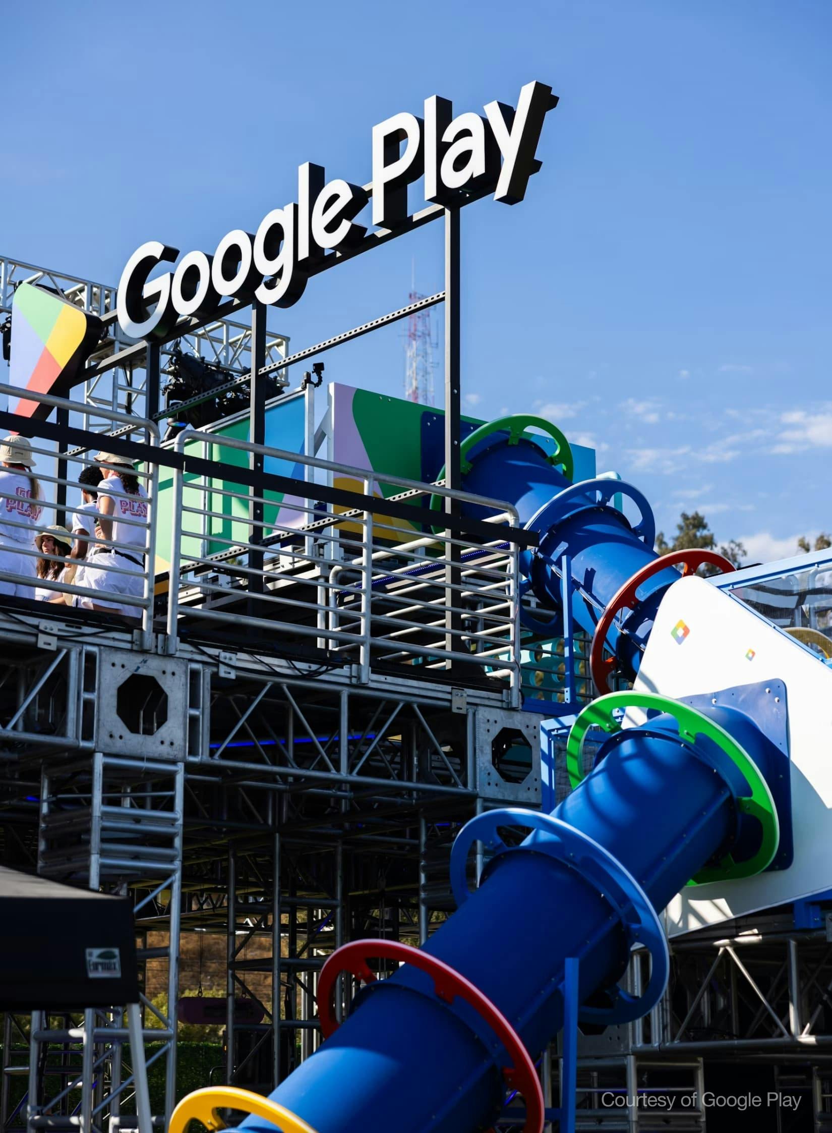 A large, multi-level Google Play outdoor interactive structure under a clear blue sky. A large white 'Google Play' sign is visible at the top. The installation includes metal truss framework, platforms with people wearing 'Go Play' shirts, and a long, blue enclosed tube slide adorned with bright red, green, and yellow rings. 'Courtesy of Google Play' is watermarked in the bottom right.