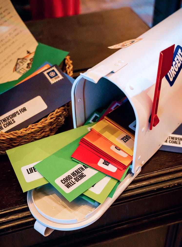 A close-up shot of a white mailbox with its door open, filled with brightly colored envelopes. The envelopes, in shades of green, red, yellow, and blue, have labels corresponding to UN Global Goals, such as 'GOOD HEALTH AND WELL-BEING' and 'PARTNERSHIPS FOR THE GOALS.' A red flag is raised on the mailbox, and a wicker basket with more mail sits beside it.