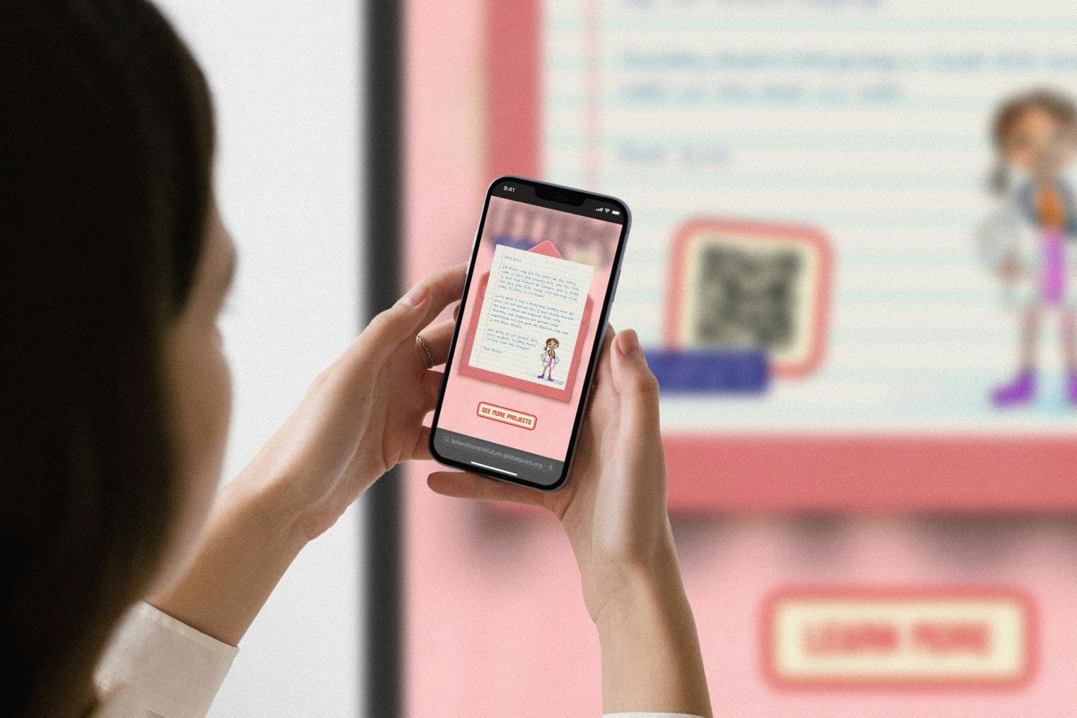 Over-the-shoulder view of a person holding a smartphone. On the phone screen is a digital letter inside a pink envelope, with a 'See More Projects' button. In the background, a large, blurred display shows the same letter with a large QR code, indicating the user has just scanned it.