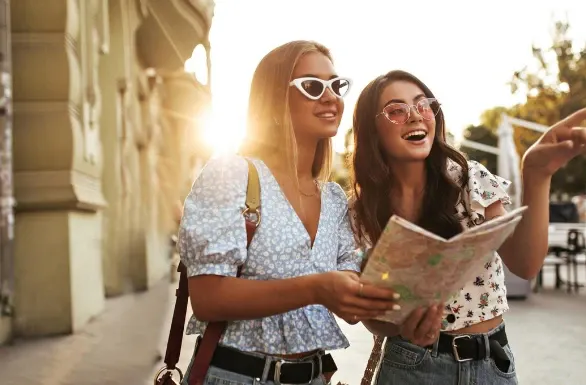 Duas amigas em um passeio ao ar livre, usando óculos de sol e roupas leves, enquanto uma delas mostra um mapa e a outra sorri em um dia ensolarado.