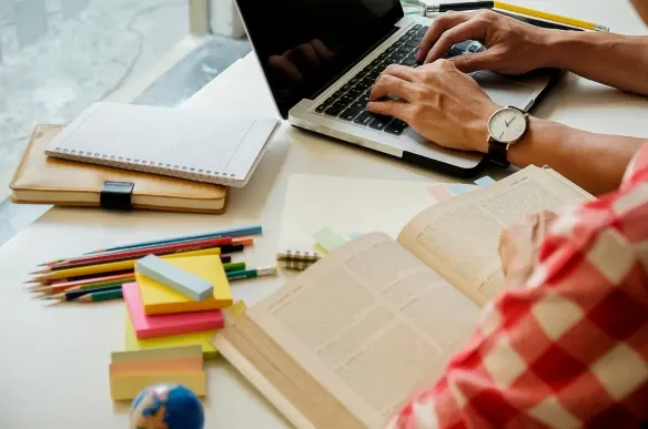 Mãos digitando em notebook em uma mesa de estudo com cadernos, lápis e post-its coloridos, ao lado de um relógio e livros abertos.