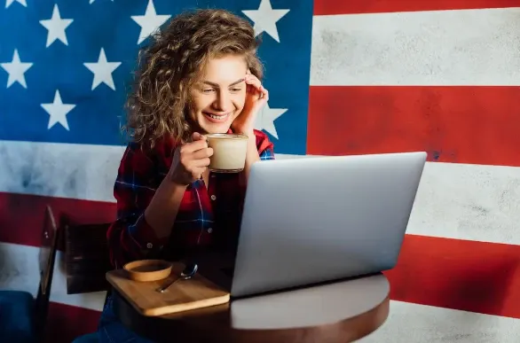 Jovem sorridente com xícara de café e notebook em mesa de madeira, em um ambiente decorado com bandeira dos Estados Unidos ao fundo