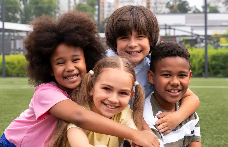 Foto ao ar livre de quatro crianças sorrindo e abraçadas no gramado de um parque, em um dia ensolarado, com prédios e árvores ao fundo.