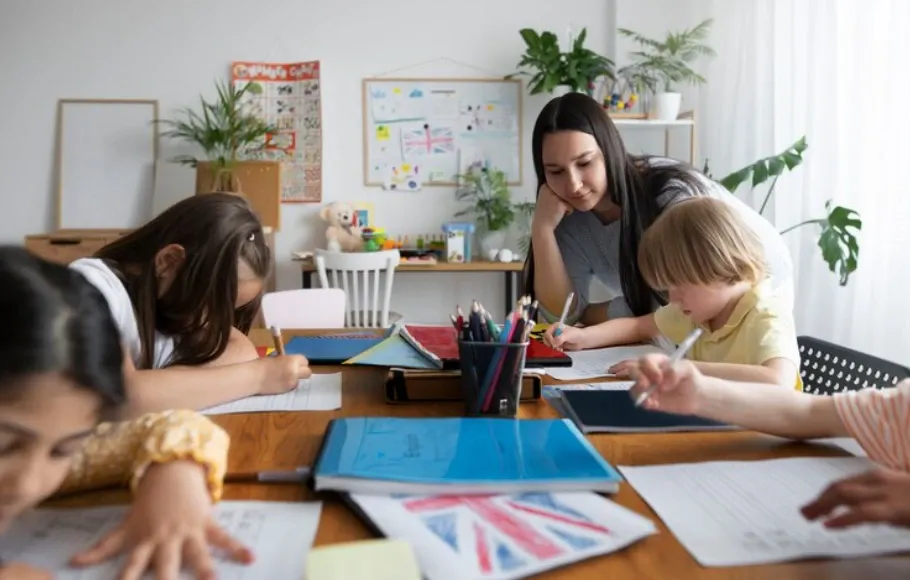 Sala de aula com uma professora ajudando crianças a desenhar e escrever em mesas de madeira, com material escolar e quadro de estudos ao fundo.