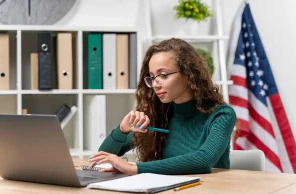 Mulher trabalhando em home office com laptop e tablet, anotando em uma mesa com prateleiras ao fundo e bandeira dos Estados Unidos ao lado
