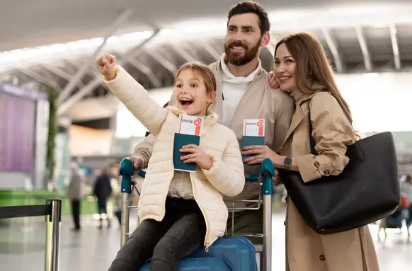 Família sorridente em estação de aeroporto, com pai, mãe e uma criança sentada em carrinho de bagagem, segurando passagens durante viagem.