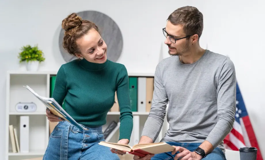 Casal de pessoas conversando e lendo um livro em um ambiente de trabalho ou estudo, com estante ao fundo e bandeira dos Estados Unidos ao lado, atmosfera amigável e colaborativa.