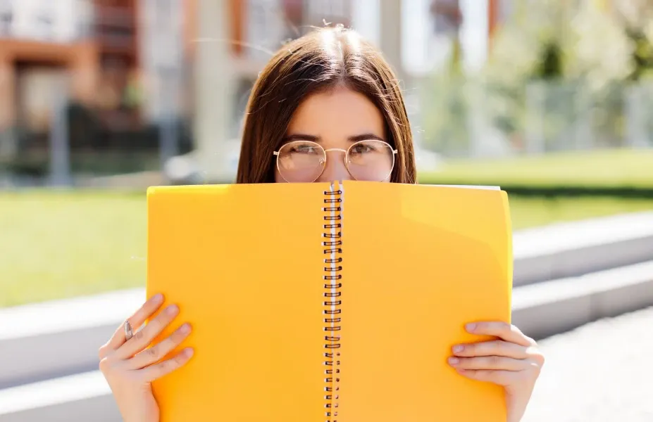 Jovem ao ar livre segurando um caderno amarelo na frente do rosto, usando óculos e luz natural em um ambiente urbano desfocado, pronta para estudar.