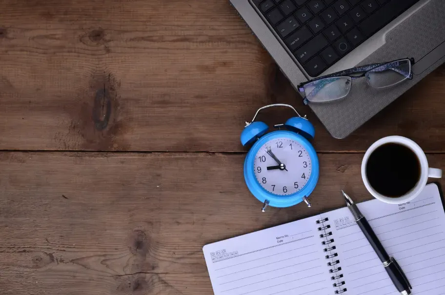 Relógio azul despertador sobre mesa de madeira, com óculos, notebook e caneca de café ao lado de caderno e caneta, sugerindo organização e rotina de trabalho
