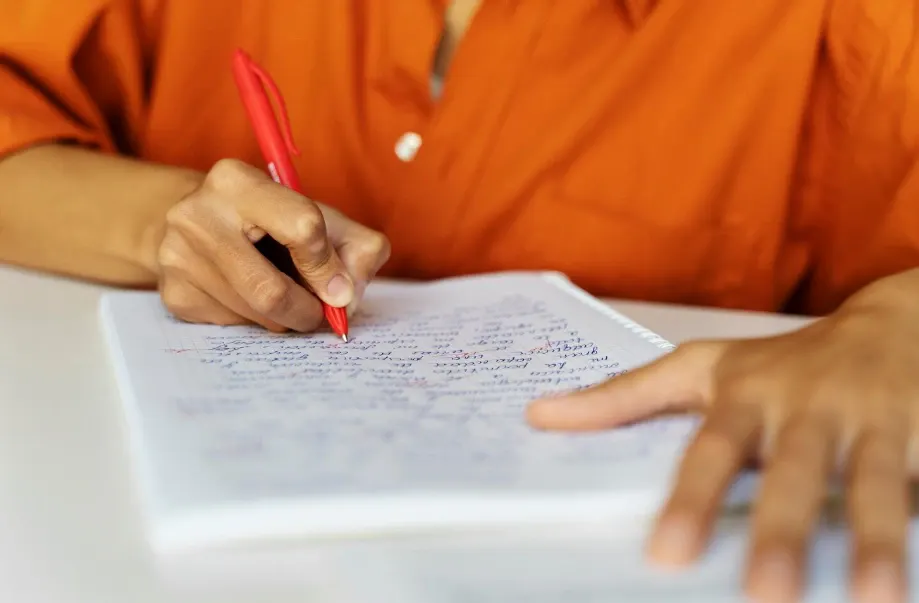 Pessoa escrevendo com caneta vermelha em uma folha de papel sobre uma mesa, com a mão apoiada no documento e foco na escrita em contexto de estudo ou trabalho