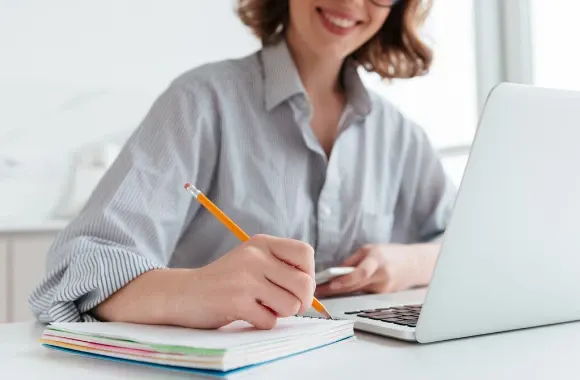 Mulher escrevendo com lápis em caderno ao lado de notebook em ambiente de escritório, com calculadora e materiais sobre a mesa