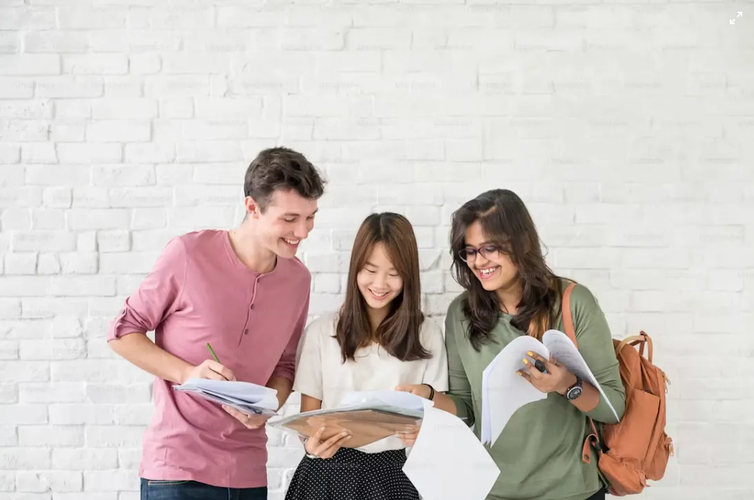 Três estudantes sorrindo e folheando livros em frente a uma parede branca, com mochila e materiais de estudo para a faculdade