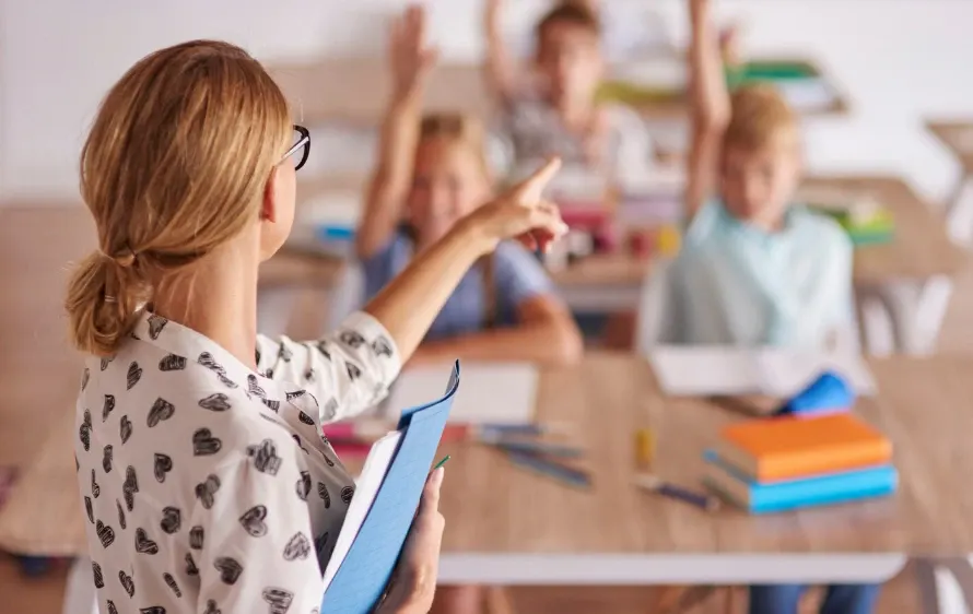Criança em primeiro plano segurando um caderno e apontando para a lousa, enquanto professores ou colegas aparecem desfocados ao fundo, em sala de aula.
