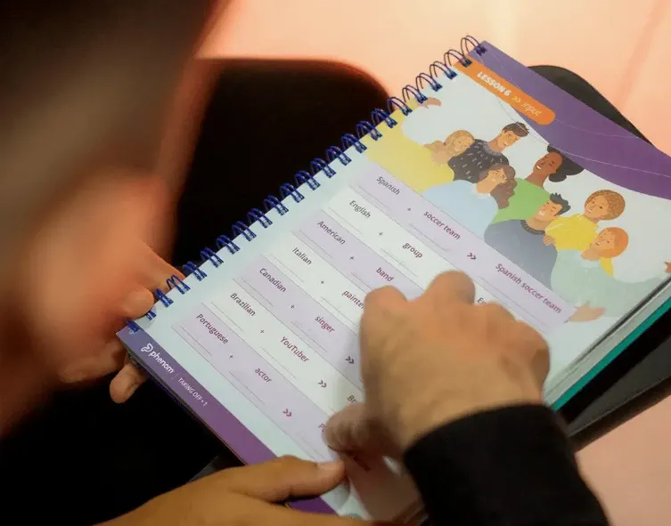 Pessoa folheando um caderno de estudos com vocabulário e exercícios de idiomas, com opções como English, Spanish e frases para aprender, em ambiente acolhedor.