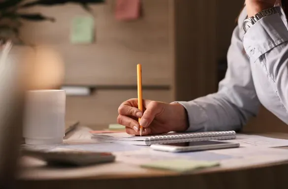 Pessoa escrevendo em um caderno com lápis amarelo em uma mesa de escritório, ao lado de bloco de notas e materiais de trabalho, em ambiente interno desfocado ao fundo