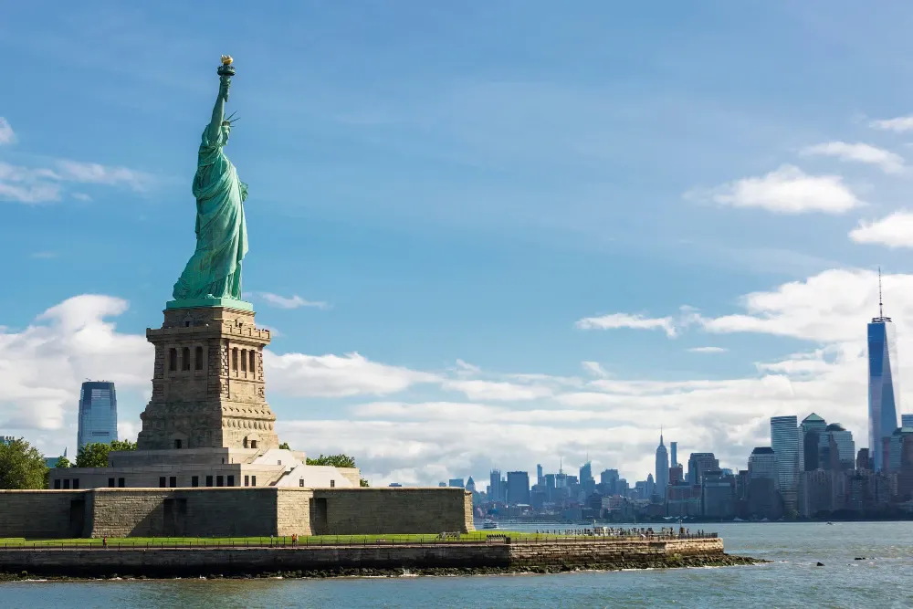 Estátua da Liberdade com o pedestal de pedra e a coroa verde, ao lado do mar, com a linha do horizonte de Manhattan e arranha-céus ao fundo sob céu azul