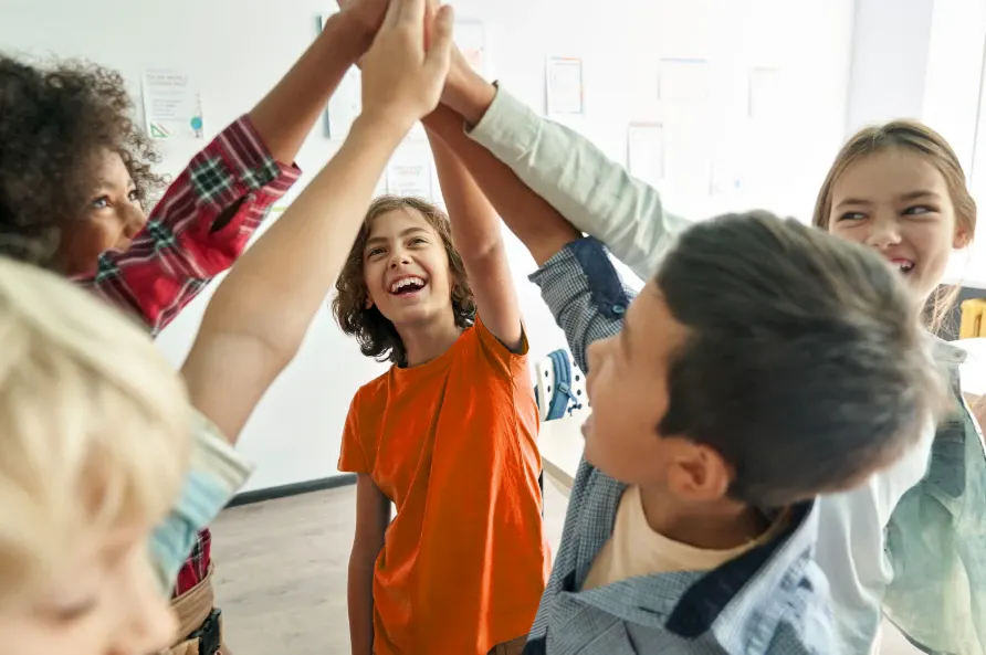 Crianças em sala de aula levantam as mãos para participar de uma brincadeira, sorrindo e brincando juntas com energia e interação no ambiente escolar.