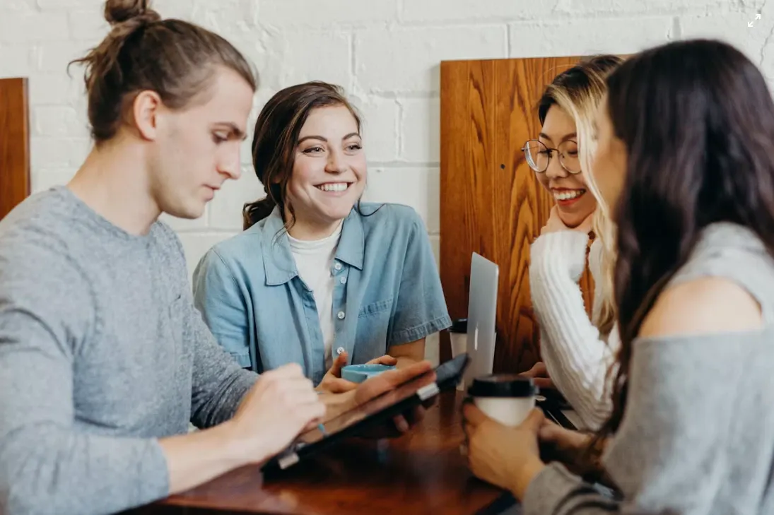 Grupo de pessoas em uma cafeteria, sorrindo e conversando enquanto compartilham um momento de trabalho e estudo ao redor de uma mesa com bebidas e celulares