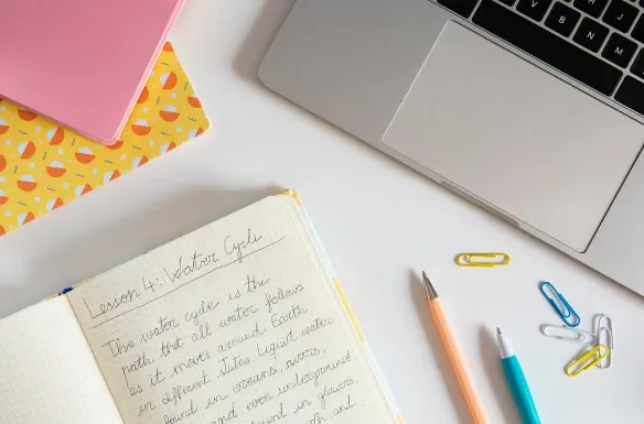 Mesa de escritório com notebook aberto, caderno quadriculado com anotações manuscritas, lápis e clipes coloridos sobre fundo claro, ao lado de um computador portátil