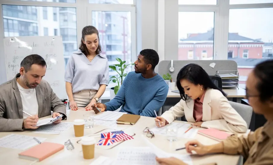 Equipe reunida em uma sala de escritório, analisando planos e discutindo um projeto em torno de uma mesa com documentos e bandeira do Reino Unido.