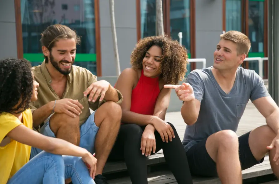 Grupo de amigos sentados em um banco ao ar livre conversando e rindo, com expressão de descontração e amizade durante um momento social.