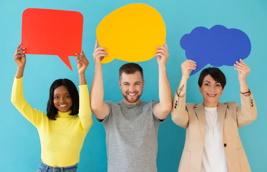 Três pessoas sorrindo seguram balões de fala coloridos redondo amarelo, nuvem azul e formas de mensagem vermelha contra um fundo azul, sugerindo comunicação e ideias.