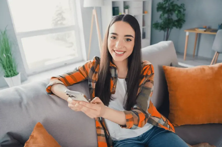 Mulher sorridente sentada no sofá da sala, usando smartphone enquanto assiste ou participa de uma chamada, com casa iluminada ao fundo e decoração moderna.