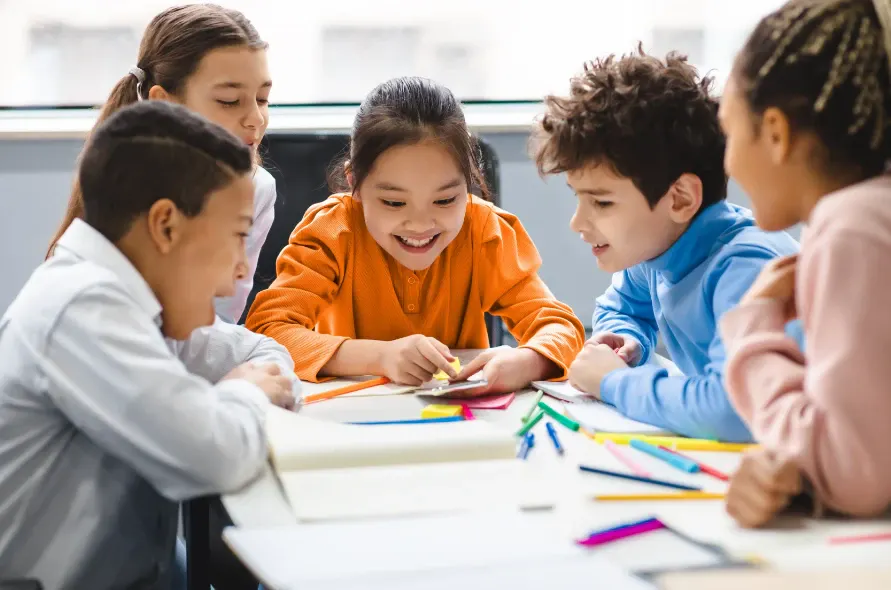 Crianças em sala de aula aprendendo juntas com materiais de desenho sobre a mesa, enquanto uma professora orienta o grupo e todos participam de atividades educativas.