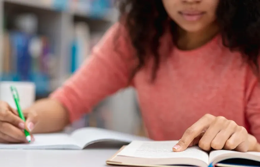 Criança sentada à mesa lendo e escrevendo em um caderno, com lápis na mão, em um ambiente escolar desfocado ao fundo.