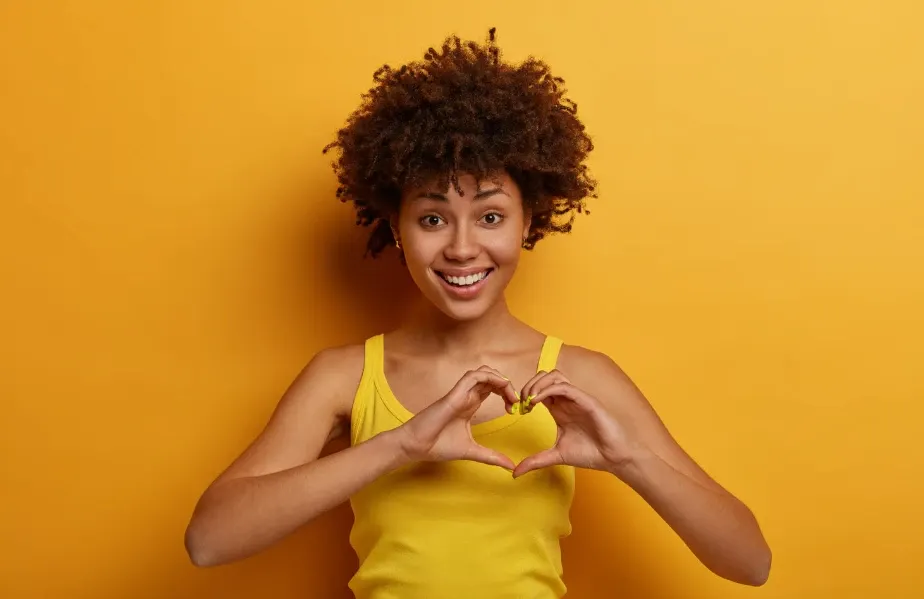 Mulher sorridente com cabelo afro encaracolado usando top amarelo em fundo laranja, formando com as mãos o símbolo de coração