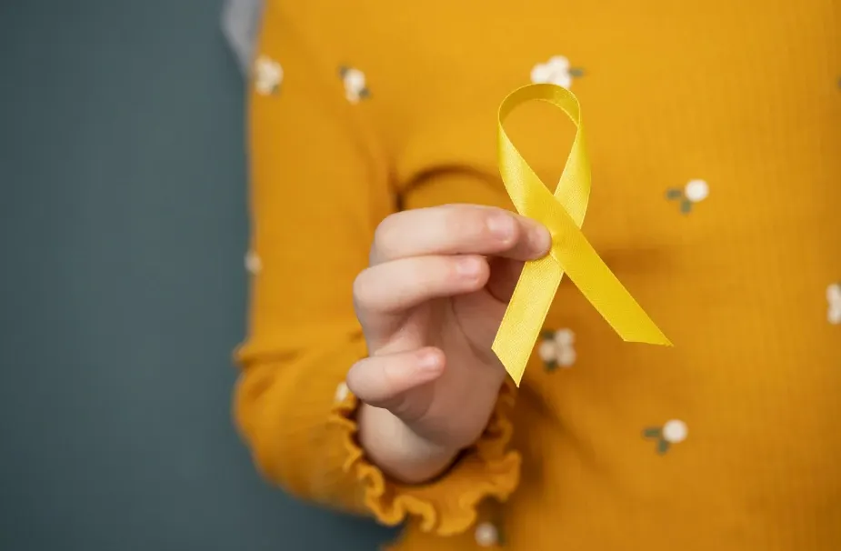 Pessoa segurando uma fita amarela laço de conscientização, com blusa amarela estampada desfocada ao fundo, em cenário de apoio à causa.