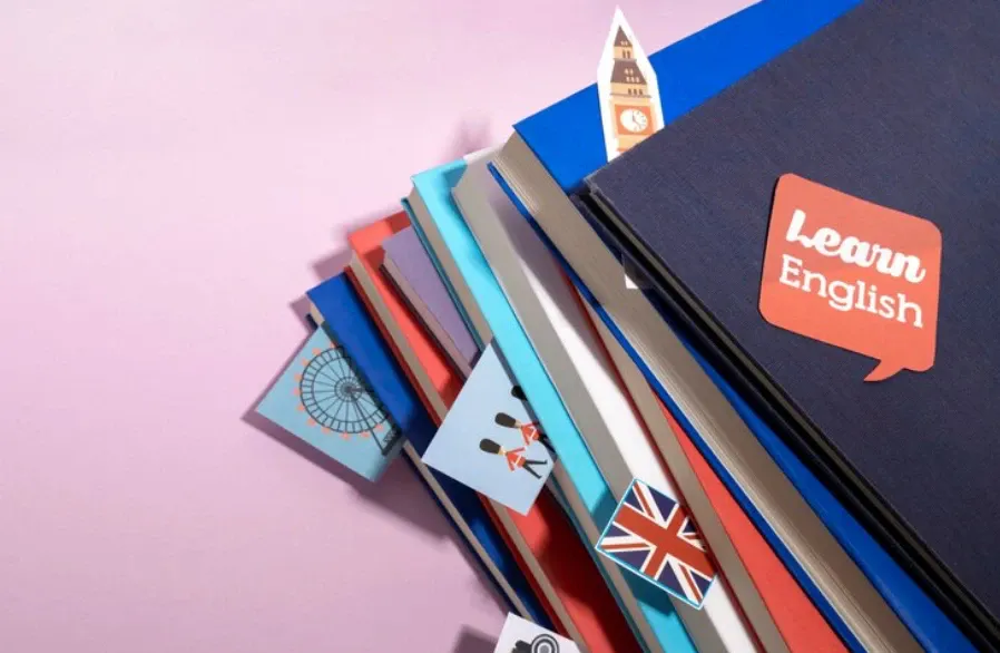 Pilha de livros de inglês com capa em azul e laranja escrito “learn English”, com marcadores e imagens de bandeiras e ícones de aprendizado ao fundo rosado.