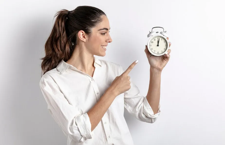 Mulher sorridente em fundo claro apontando para um relógio despertador analógico na mão, transmitindo ideia de pontualidade e tempo