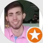 Foto de um homem sorrindo em frente a um fundo verde, com um selo circular laranja com uma estrela no canto inferior direito.