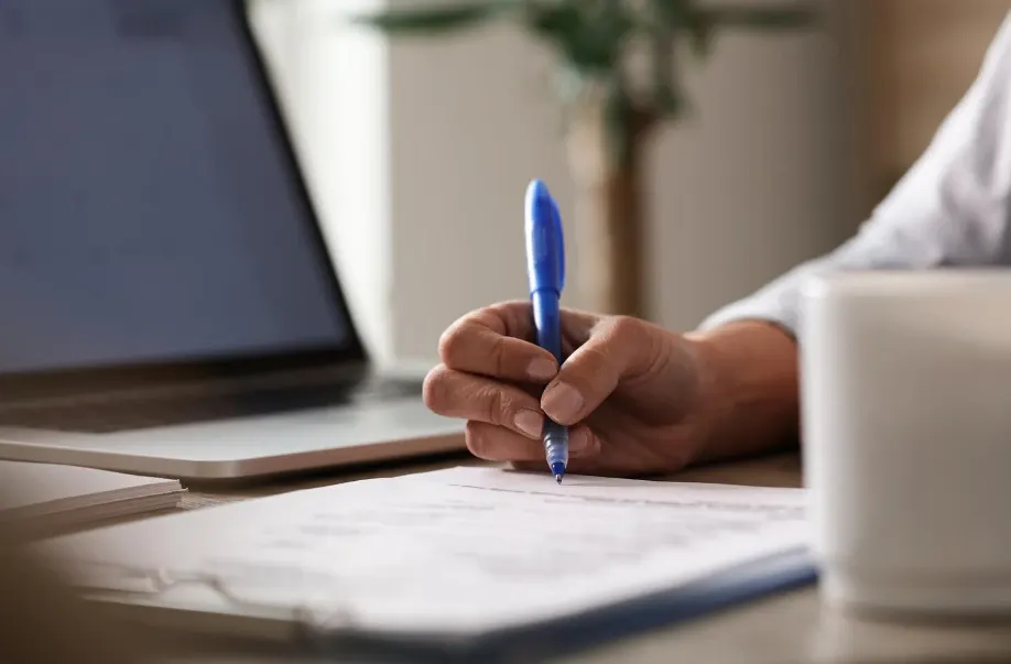 Pessoa escrevendo com caneta azul em uma folha sobre uma mesa, com notebook ao fundo, em um ambiente de escritório, planejamento e trabalho profissional