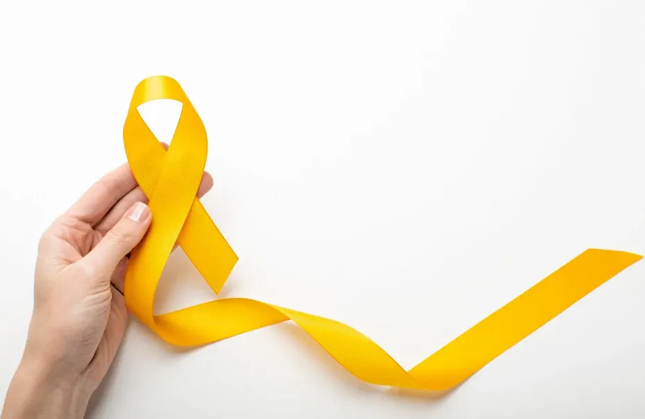 Mão segurando um laço amarelo no fundo branco, símbolo de conscientização e prevenção, com fita em destaque e espaço para texto.